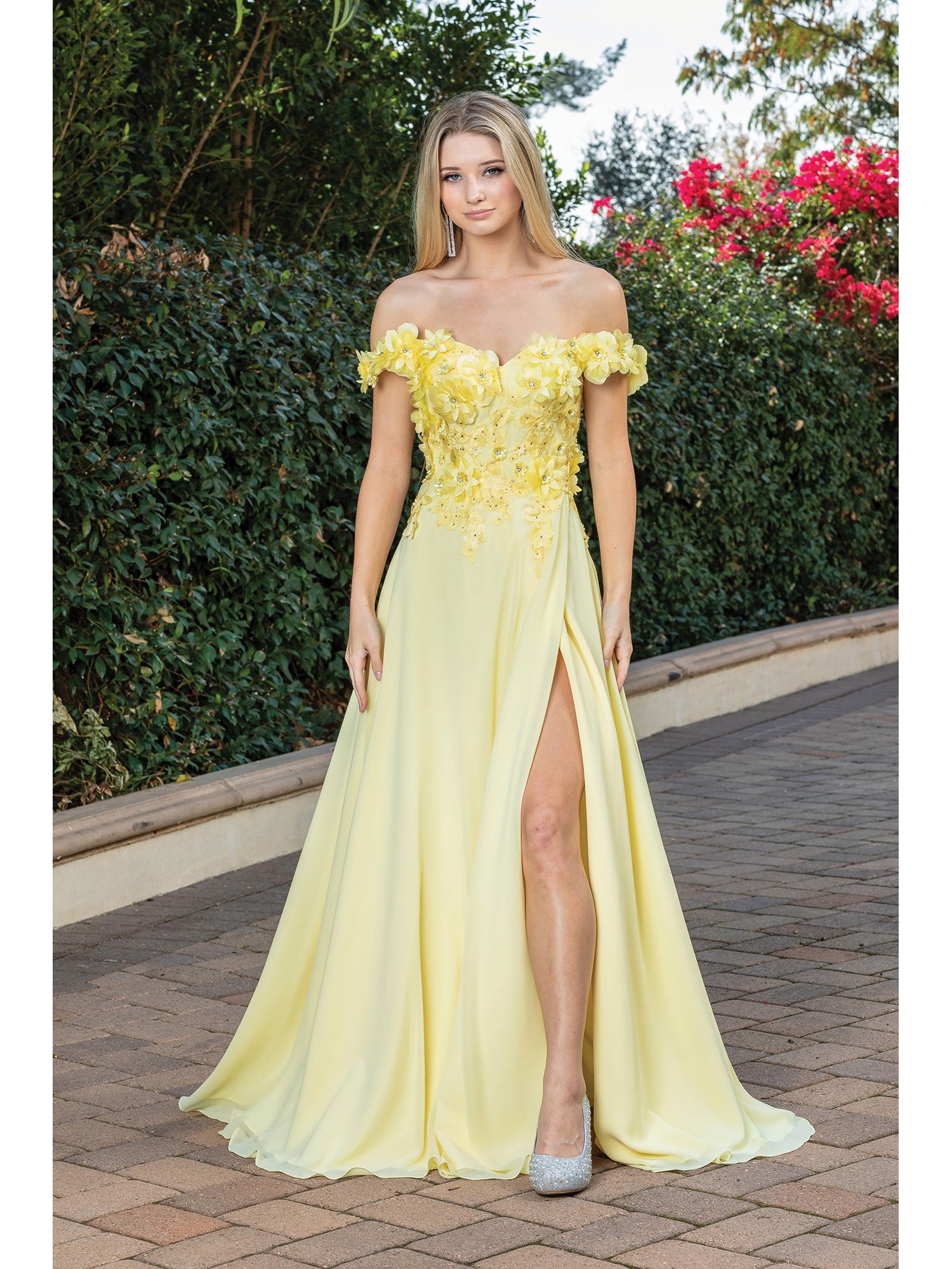 A woman stands outdoors in a Long Corset-Back Prom Dress with Beaded Bodice, featuring a light yellow color, off-the-shoulder style, thigh-high slit, and silver high heels. Shrubs and flowers are visible in the background.